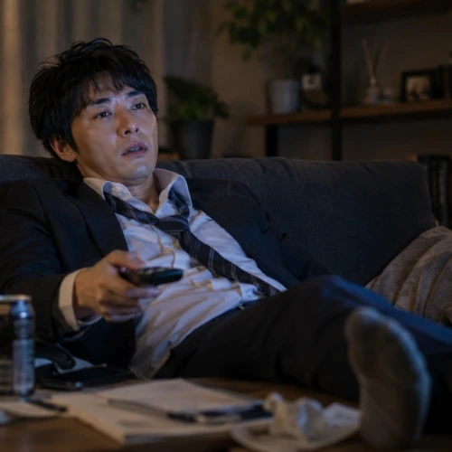 疲れて帰宅したスーツ姿の男性がソファでくつろぎながら、眠そうな表情で夢中になってドラマを見ている様子
