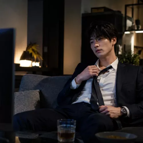 仕事から帰宅したスーツ姿の男性がネクタイを緩めながらソファでドラマを見てくつろぐ夜のリビング風景